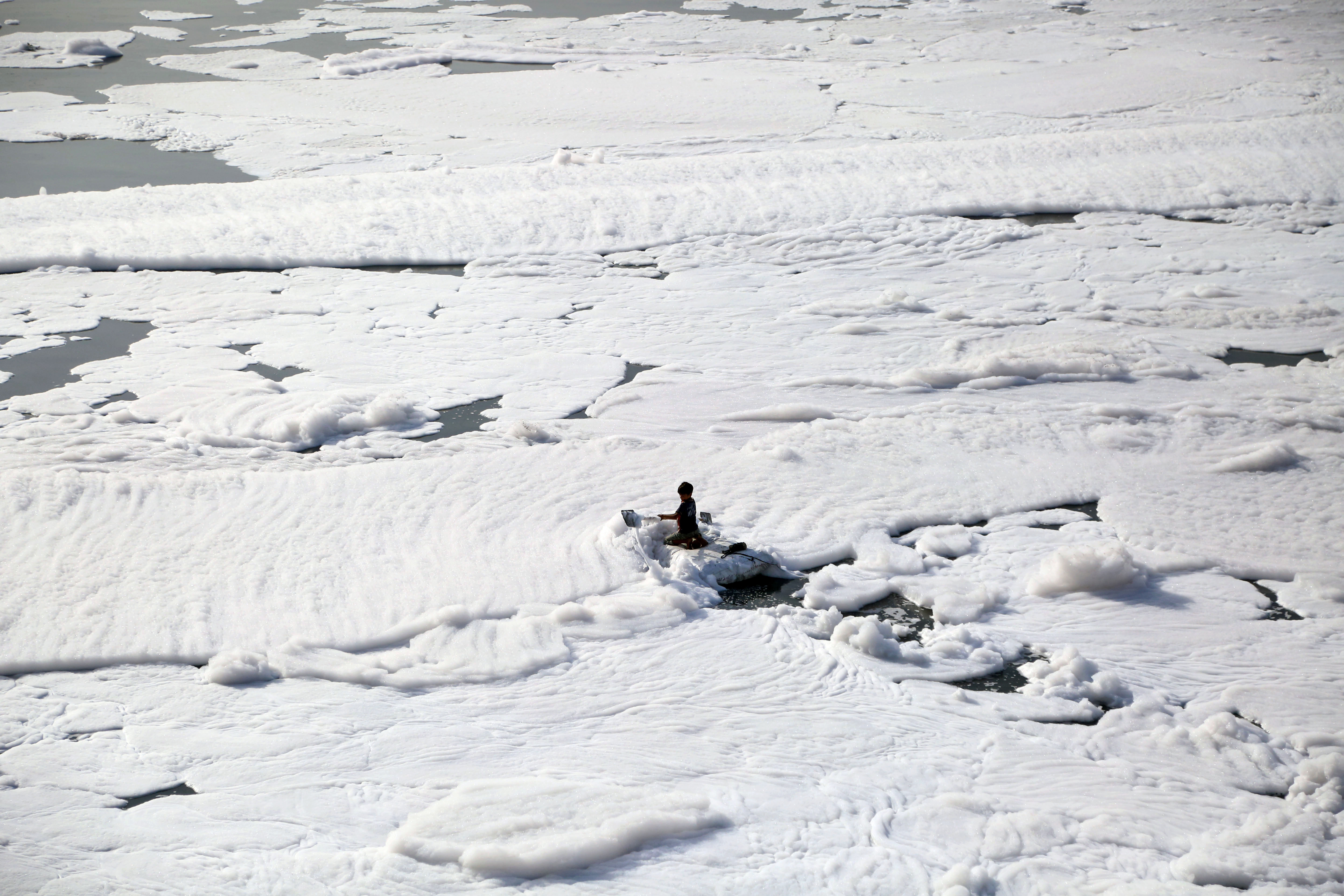 A boy rows a makeshift raft through toxic foam floating on the Yamuna river