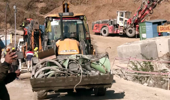 An excavator carries the damaged parts of the auger machine used for the drilling to rescue the trapped workers inside the Silkyara Tunnel